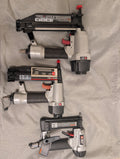 Three cordless nail guns in black, gray, and silver are arranged vertically on a beige fabric background.