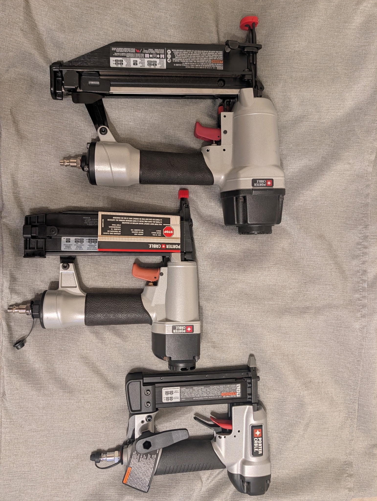 Three cordless nail guns in black, gray, and silver are arranged vertically on a beige fabric background.