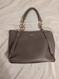 A taupe brown leather handbag with gold-tone chain handles and a gold 'ANNE KLEIN' logo on the front, laid flat on a light gray fabric background.