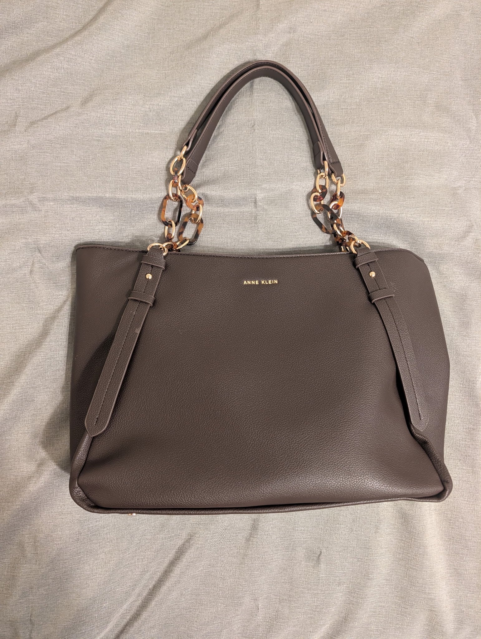 A taupe brown leather handbag with gold-tone chain handles and a gold 'ANNE KLEIN' logo on the front, laid flat on a light gray fabric background.