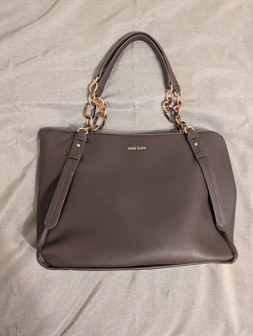 A taupe brown leather handbag with gold-tone chain handles and a gold 'ANNE KLEIN' logo on the front, laid flat on a light gray fabric background.