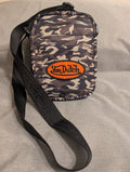 A small crossbody bag with a dark camouflage pattern, featuring a prominent orange oval patch with the brand name 'Von Dutch' and a black adjustable shoulder strap, laid flat on a gray fabric surface.