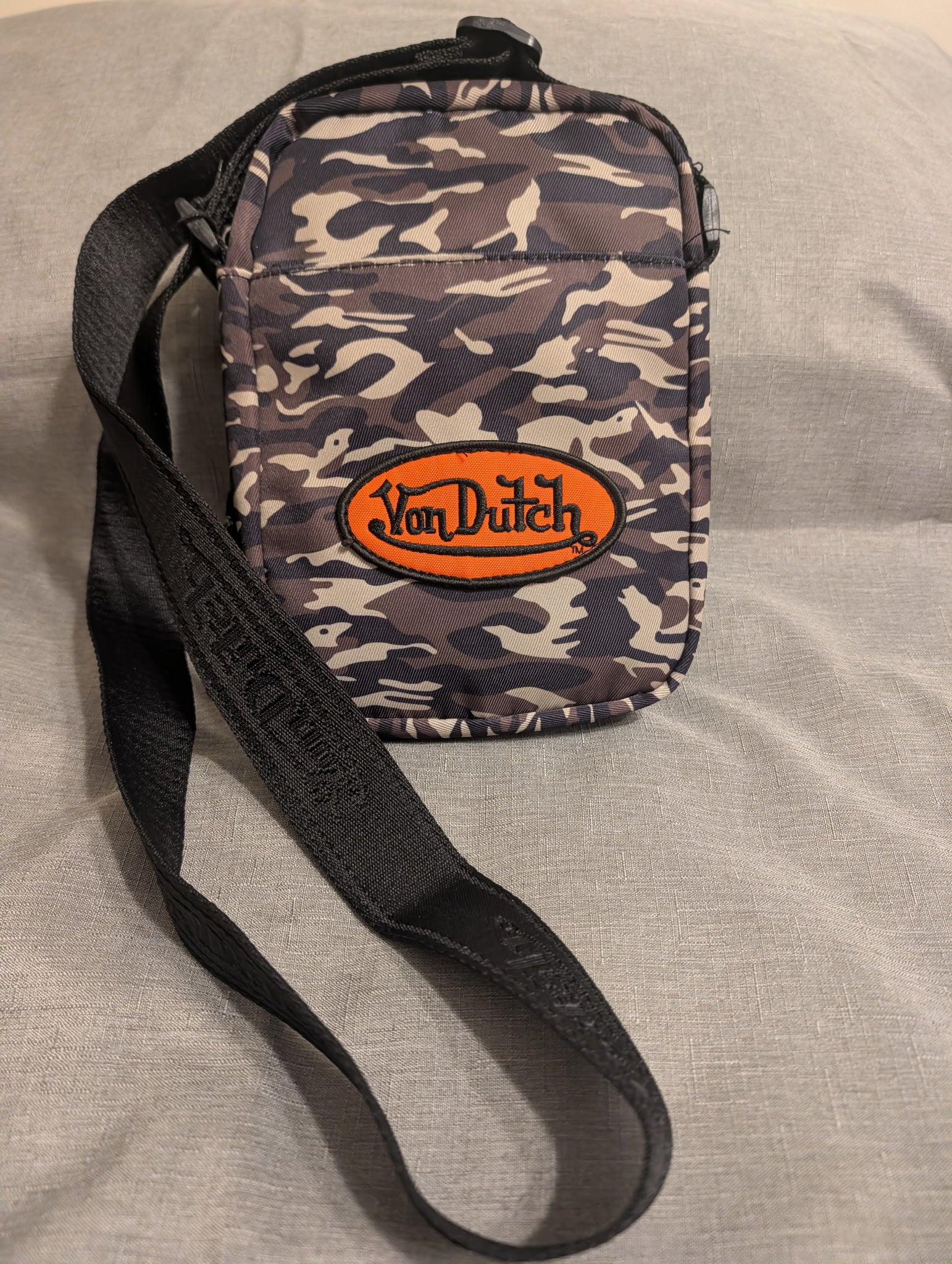 A small crossbody bag with a dark camouflage pattern, featuring a prominent orange oval patch with the brand name 'Von Dutch' and a black adjustable shoulder strap, laid flat on a gray fabric surface.