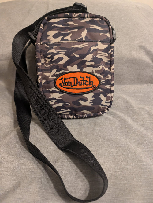 A small crossbody bag with a dark camouflage pattern, featuring a prominent orange oval patch with the brand name 'Von Dutch' and a black adjustable shoulder strap, laid flat on a gray fabric surface.