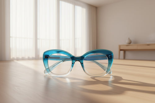 A pair of chunky, oversized cat-eye style eyeglasses with a translucent blue and clear plastic frame, resting on a dark textured carpet. A small blue sticker is visible on the lower right lens.