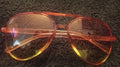 A pair of aviator-style sunglasses with translucent orange frames and yellow-tinted lenses, resting on a dark textured carpet.