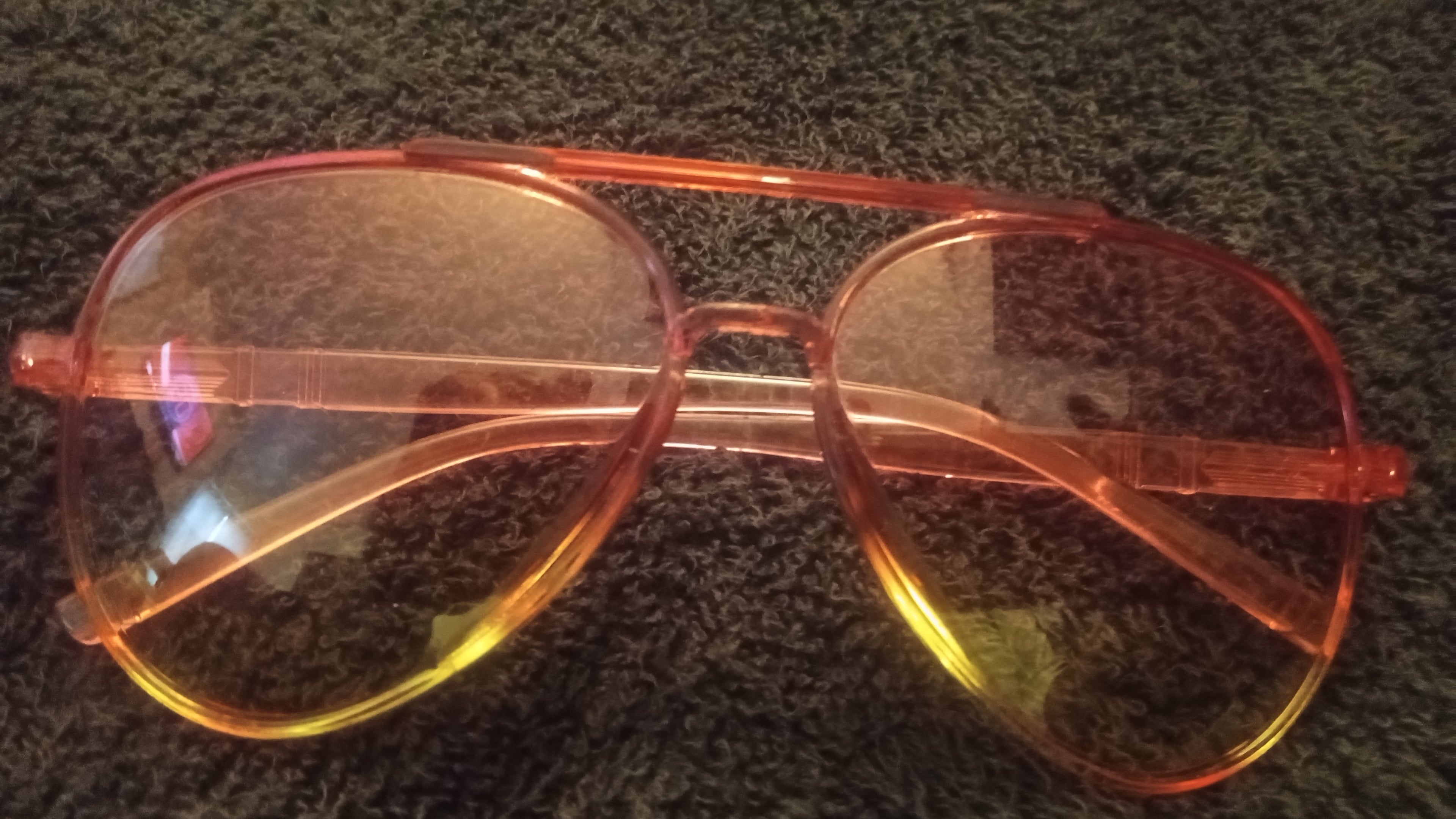 A pair of aviator-style sunglasses with translucent orange frames and yellow-tinted lenses, resting on a dark textured carpet.