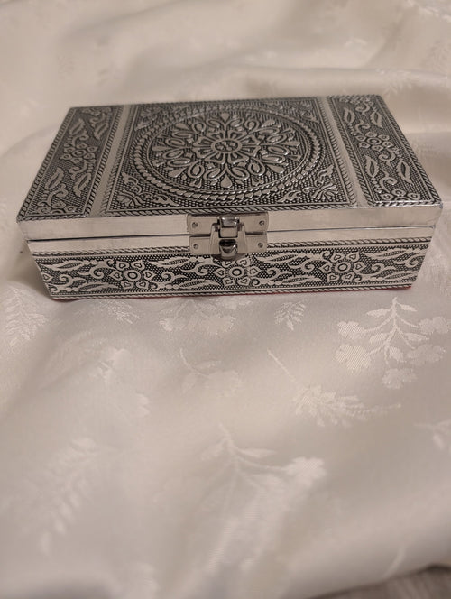 An ornate rectangular silver-toned metal box with intricate floral and geometric engravings on the lid and sides, featuring a central medallion and a latch closure, resting on a white patterned fabric surface.
